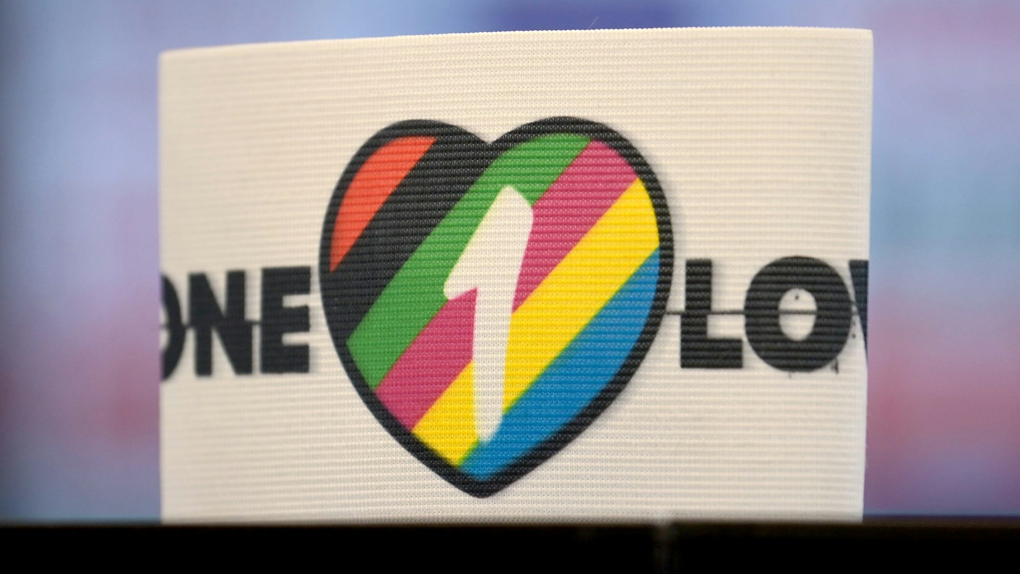Eine Armbinde, die ein Herz und die Aufschrift „One Love“ trägt, steht auf einer Oberfläche. Das Herz ist mit bunten Streifen sowie der Zahl 1 gefüllt. Die Farben symbolisieren die Vielfalt unterschiedlicher Lebensentwürfe.