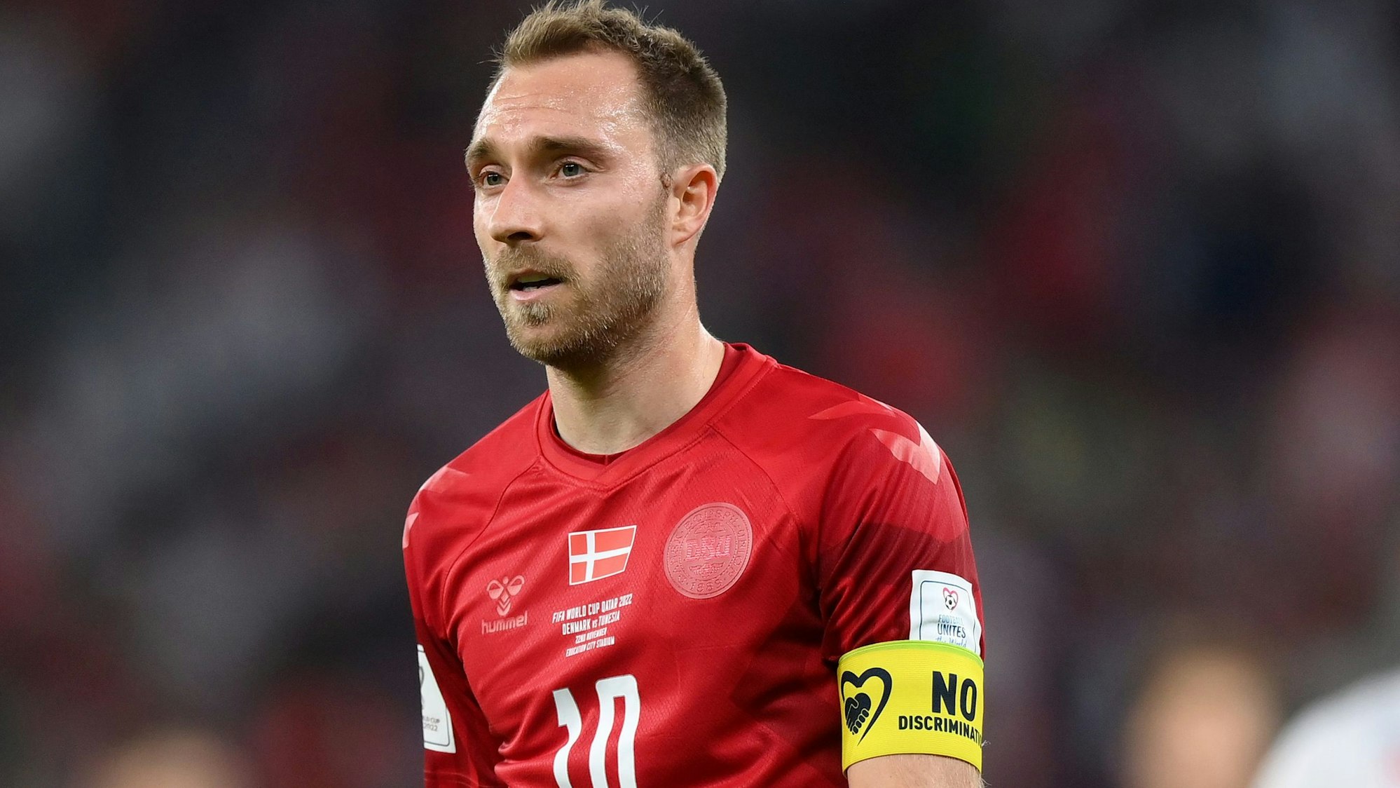 Der dänische Fußballspieler Christian Eriksen trägt am linken Oberarm eine gelbe Binde, auf der in schwarzer Schrift „No Discrimination“ sowie ein schwarz umrissenes Herz abgebildet ist.