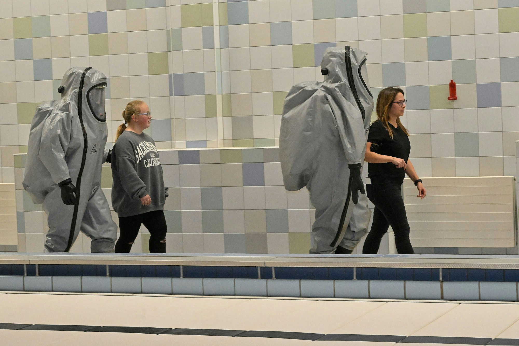 Zwei Feuerwehrleute in in grauen Schutzanzügen bringen zwei Schülerinnen aus der Schwimmhalle in Sicherheit.