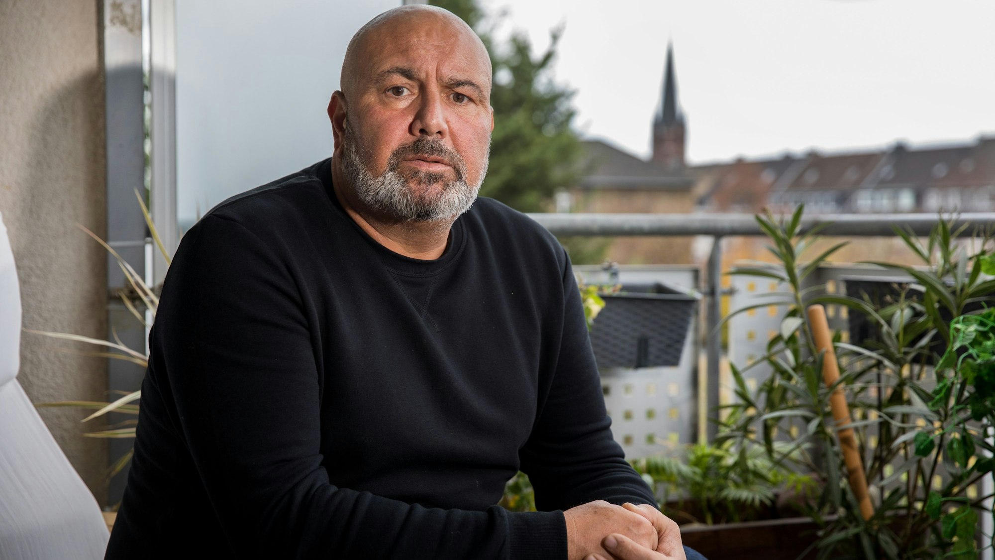 Ersin Seyhun sitzt auf einem Sessel auf seinem Balkon, im Hintergrund ragt ein Kirchturm in den grauen Himmel.