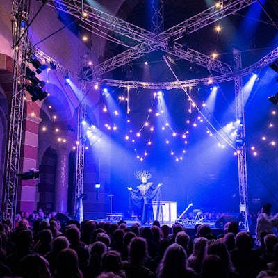 In der Kirche wurde eine Bühne aufgebaut auf der eine Sängerin im Kostüm steht. Vor der Bühne sitzen Zuschauer. Das Licht ist blau.