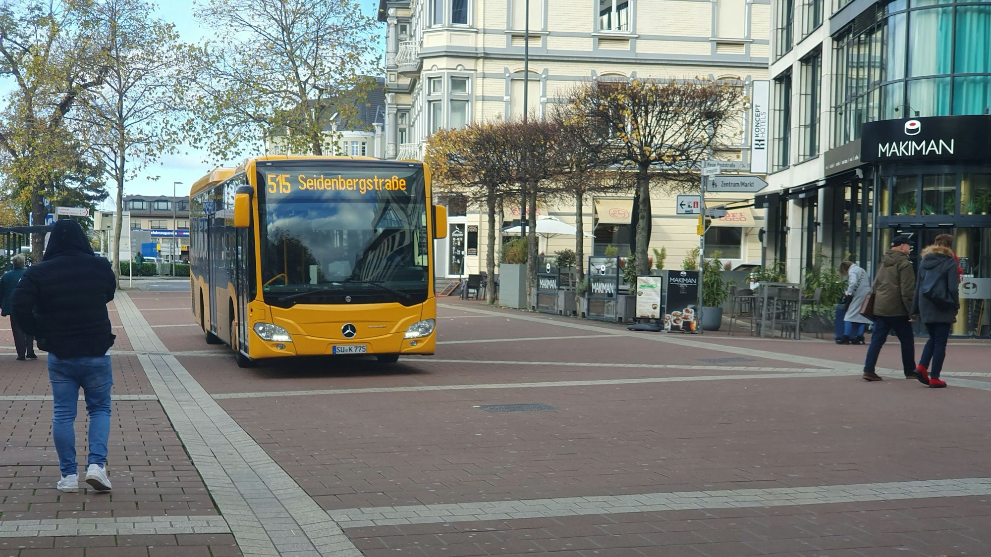 Ein Linienbus der Rhein-Sieg-Verkehrsgesellschaft (RSVG) fährt über eine gepflasterte Straße, die von Bäumen gesäumt ist. Passanten gehen an beiden Seiten.