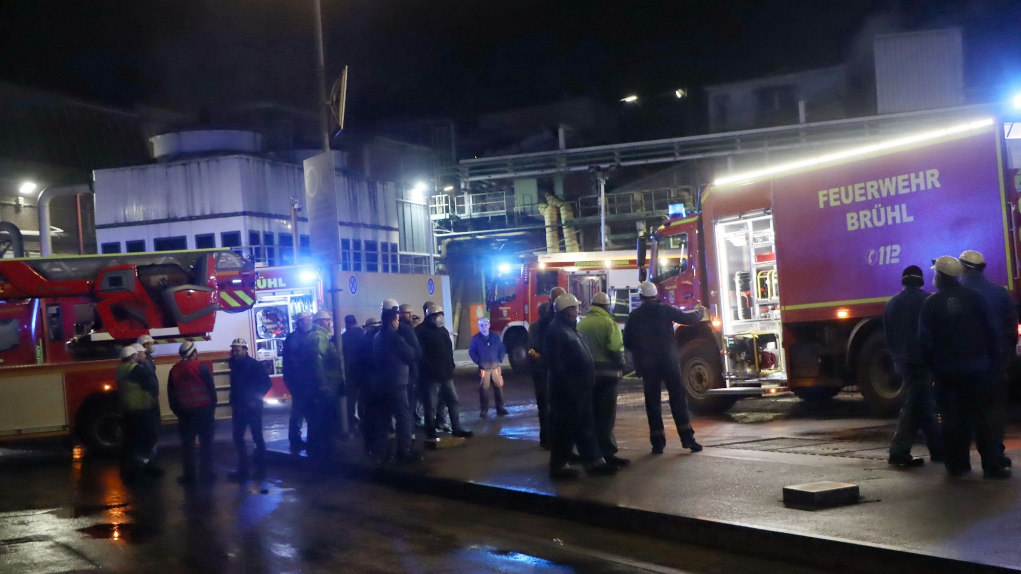 Einige Menschen stehen in der Nacht vor dem Brühler Eisenwerk zwischen Feuerwehrwagen. Fast alle tragen Schutzhelme.