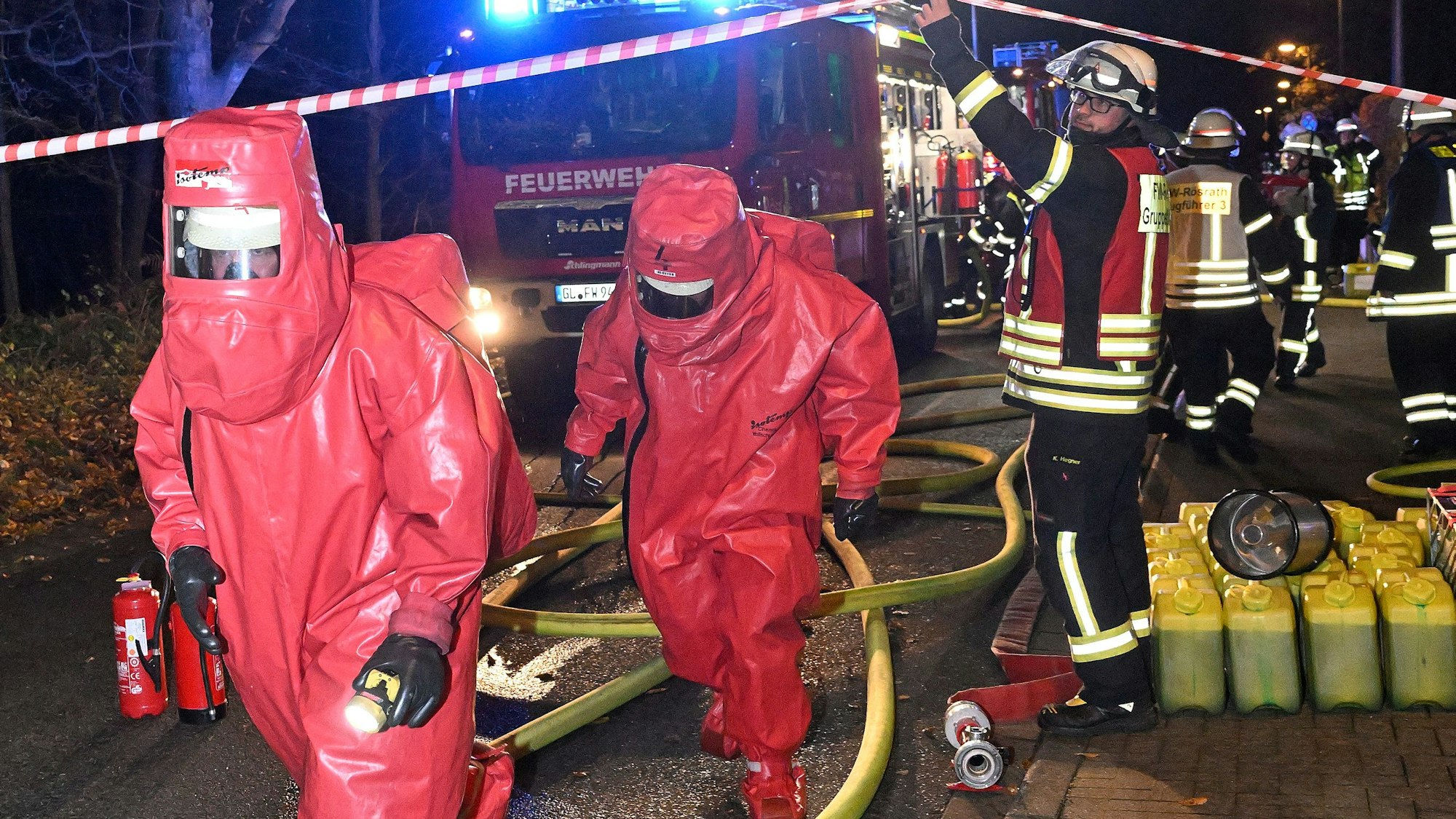 Feuerwehrleute in roten Chemikalienschutzanzügen gehen unter einer Absperrung hindurch.
