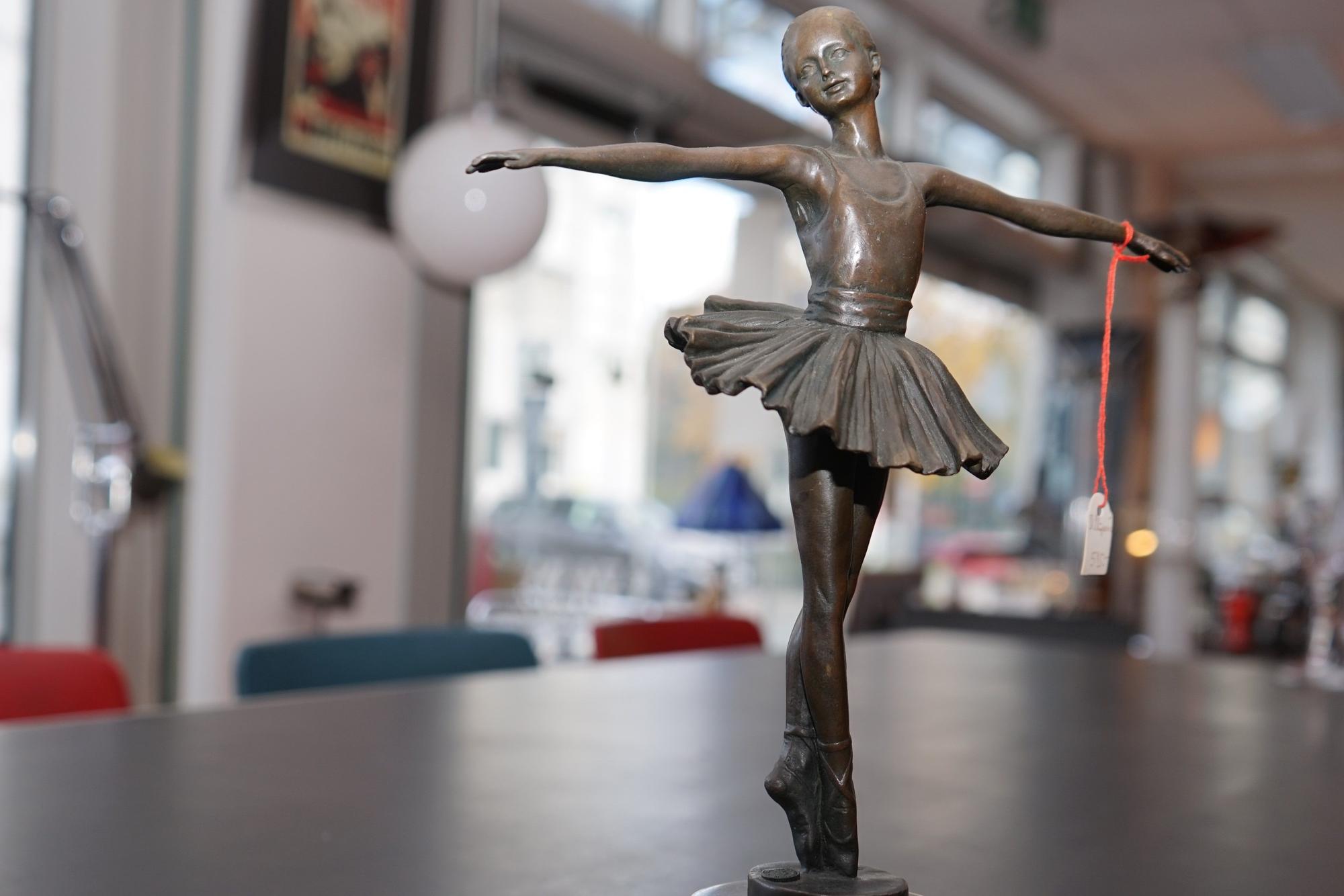 Eine Ballerina aus Metall steht auf einem Holztisch in Norbert Essers Antiquariat in Bergisch Gladbach. Sie hat ihre Arme ausgebreitet und trägt ein Tutu.