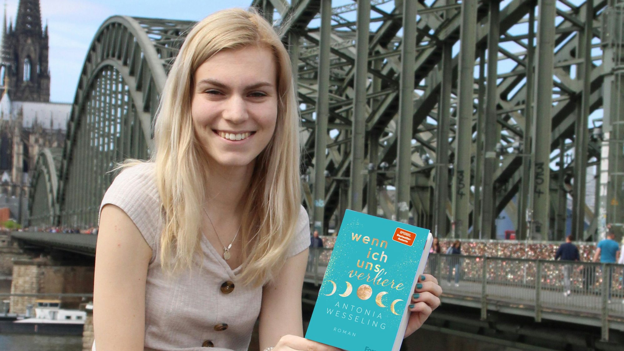 Antonia Wesseling sitzt am Rhein auf einer Mauer, hinter ihr die Hohenzollernbrücke und der Dom. In den Händen hält sie ihren Roman "Wenn ich uns verliere".