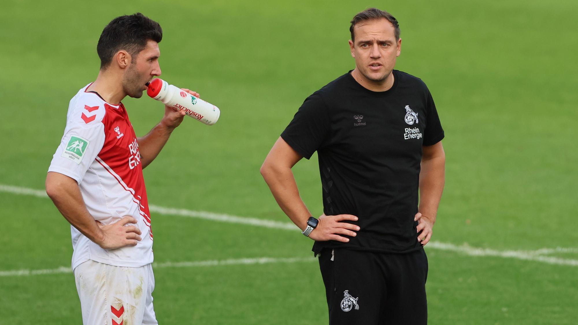 Ein Fußballer und sein Trainer stehen während einer Pause auf dem Spielfeld. Der Spieler trinkt aus einer Flasche.