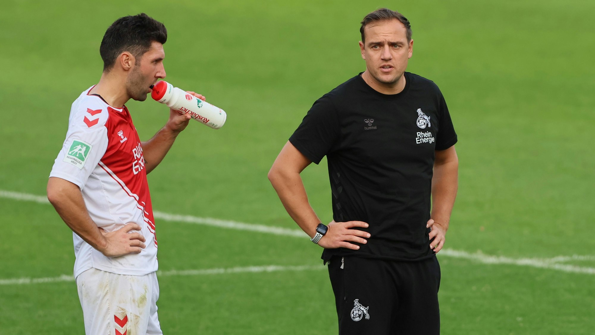 Ein Fußballer und sein Trainer stehen während einer Pause auf dem Spielfeld. Der Spieler trinkt aus einer Flasche.