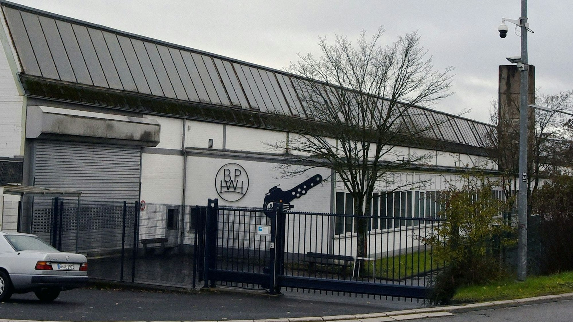 Ein längliches, weißes Gebäude ist hinter einem schwarzen Zaun mit Tor zu sehen. Am Gebäude hängt ein das Firmen-Logo BPW.