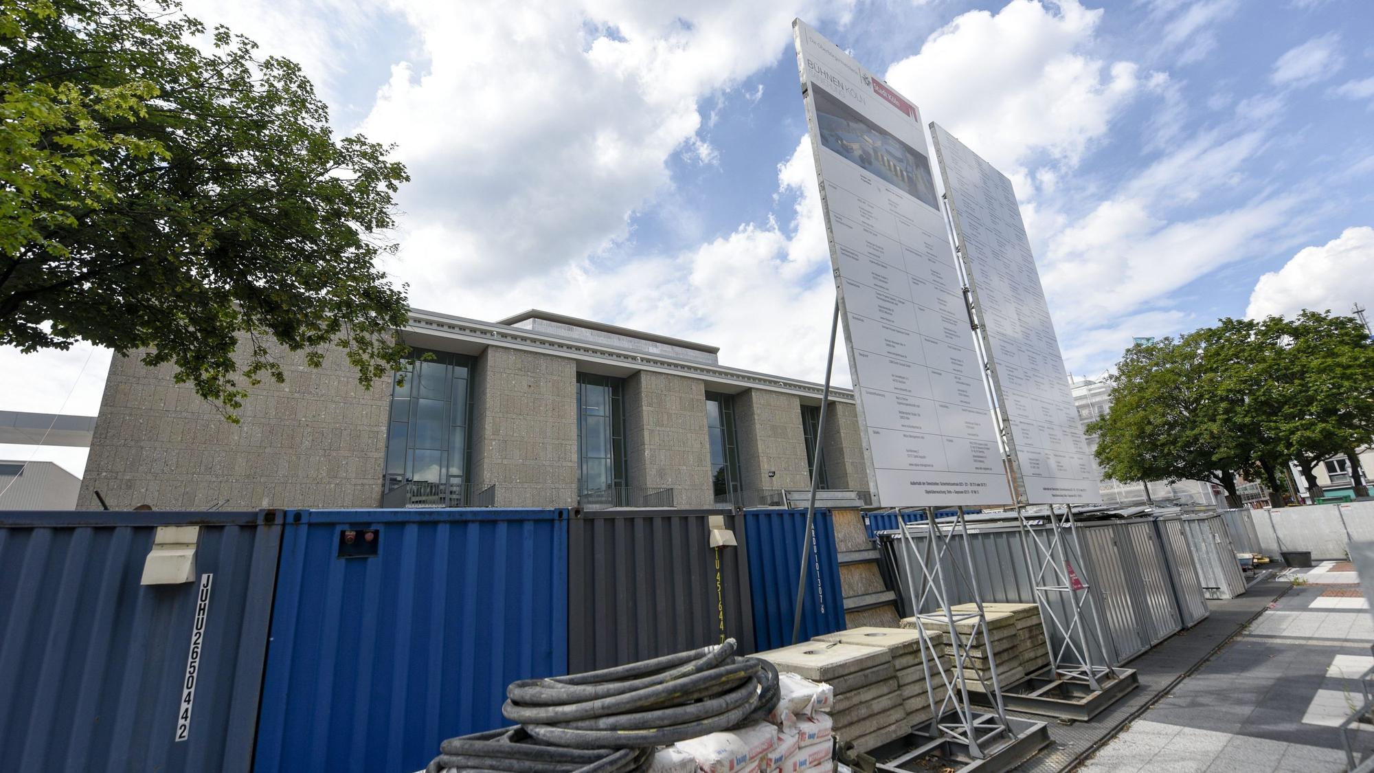 Das Kölner Operngebäude wird zum Teil von einem blauen Metall-Bauzaun verdeckt, vor dem Baumaterialien lagern. Davor steht ein Schild, auf dem die an der Baumaßnahme beteiligten Firmen aufgeführt sind.