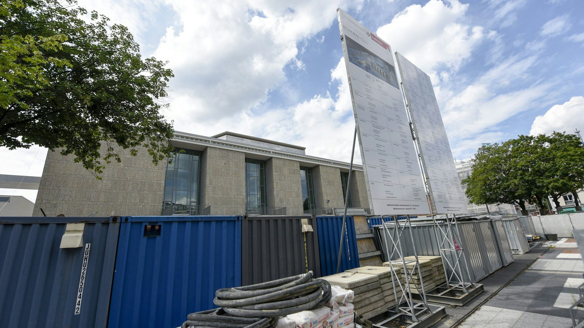Das Kölner Operngebäude wird zum Teil von einem blauen Metall-Bauzaun verdeckt, vor dem Baumaterialien lagern. Davor steht ein Schild, auf dem die an der Baumaßnahme beteiligten Firmen aufgeführt sind.