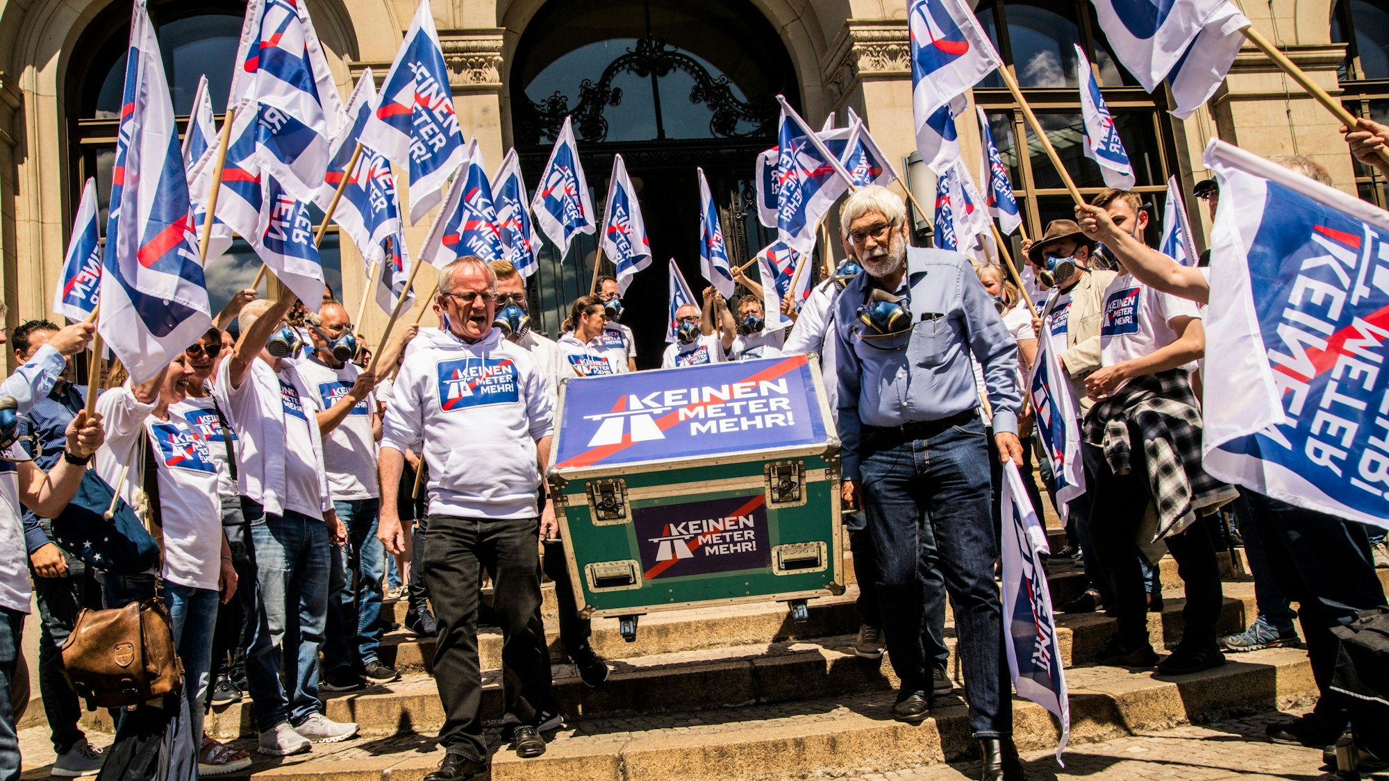 Auf einer Treppe stehen mehrere Dutzend Menschen, sie tragen Pullover und schwenken Fahnen mit der Aufschrift „Keinen Meter mehr!“