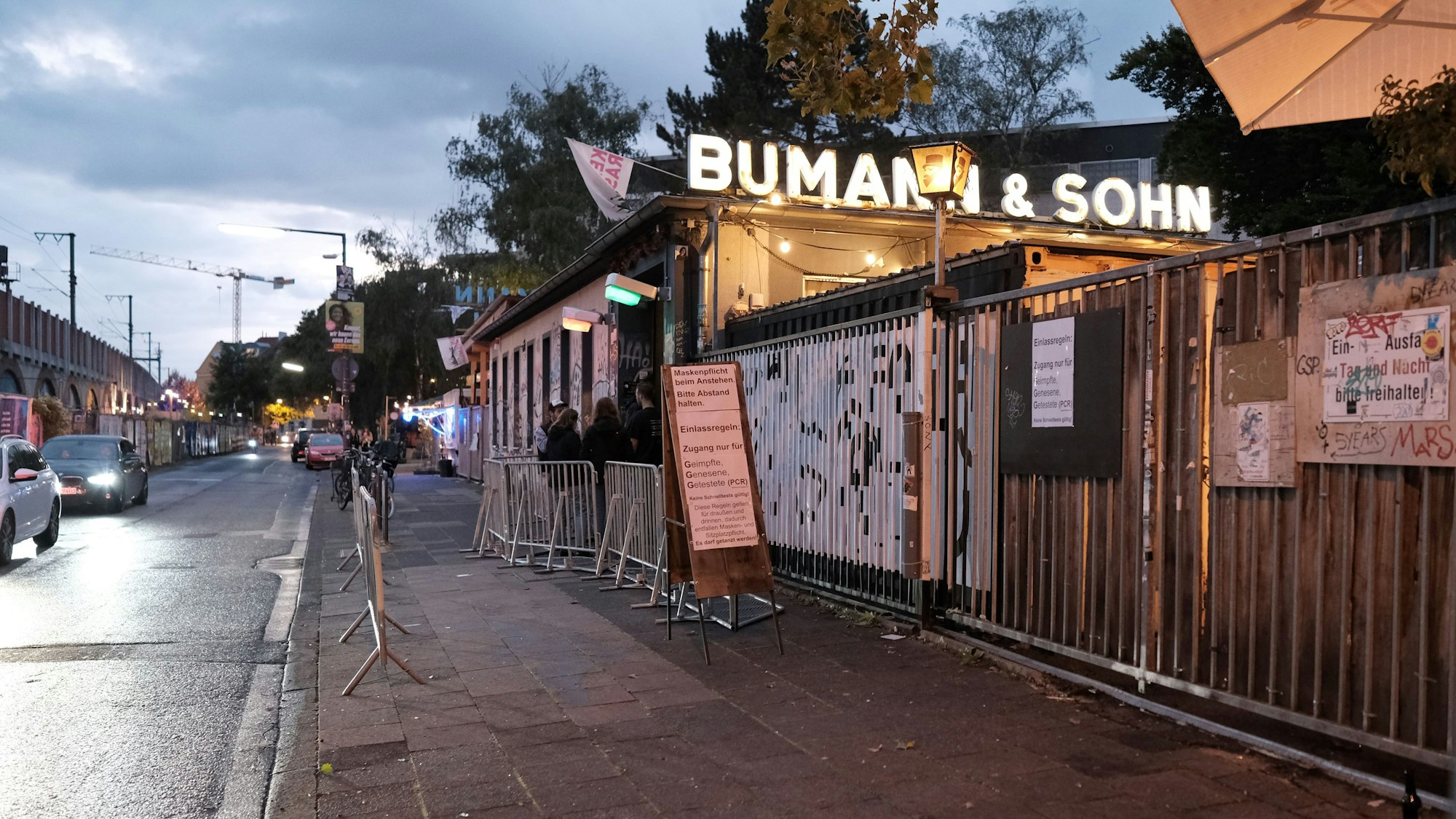 Außenaufnahmen vor dem Bumann und Sohn, links fahren Autos, rechts oben ist der leuchtende Schriftzug der Bar zu sehen, davor stehen Menschen an, um in den Laden zu kommen.