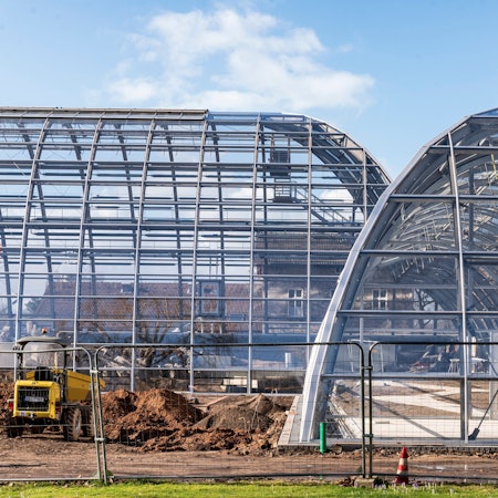 Neubau der Schaugewächshäuser und der Orangerie im Botanischen Garten. Zwei Schaugewächshäuser ohne Pflanzen darin, vorne ein Bauzaun mit einem Bagger.
