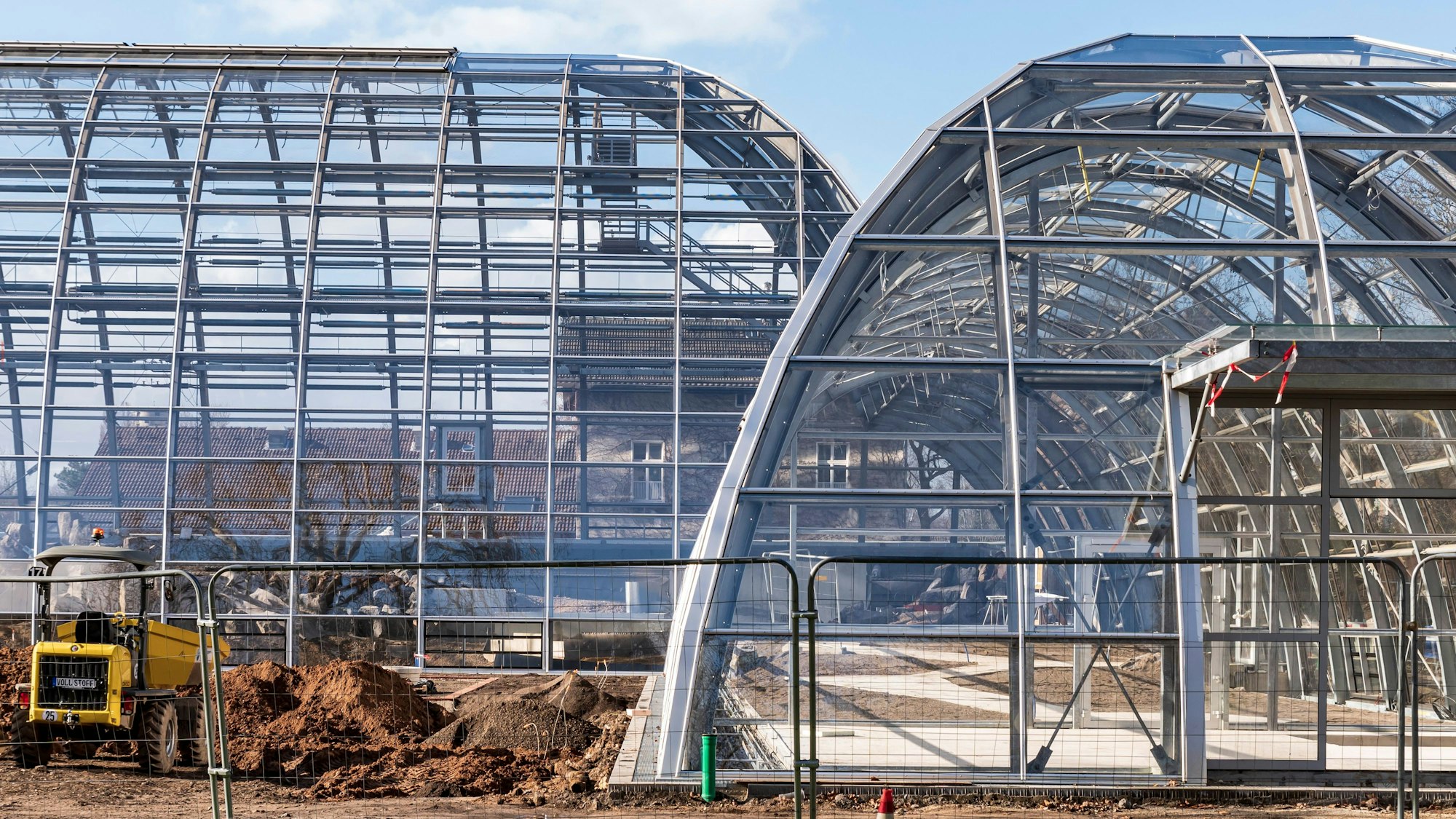 Neubau der Schaugewächshäuser und der Orangerie im Botanischen Garten. Zwei Schaugewächshäuser ohne Pflanzen darin, vorne ein Bauzaun mit einem Bagger.