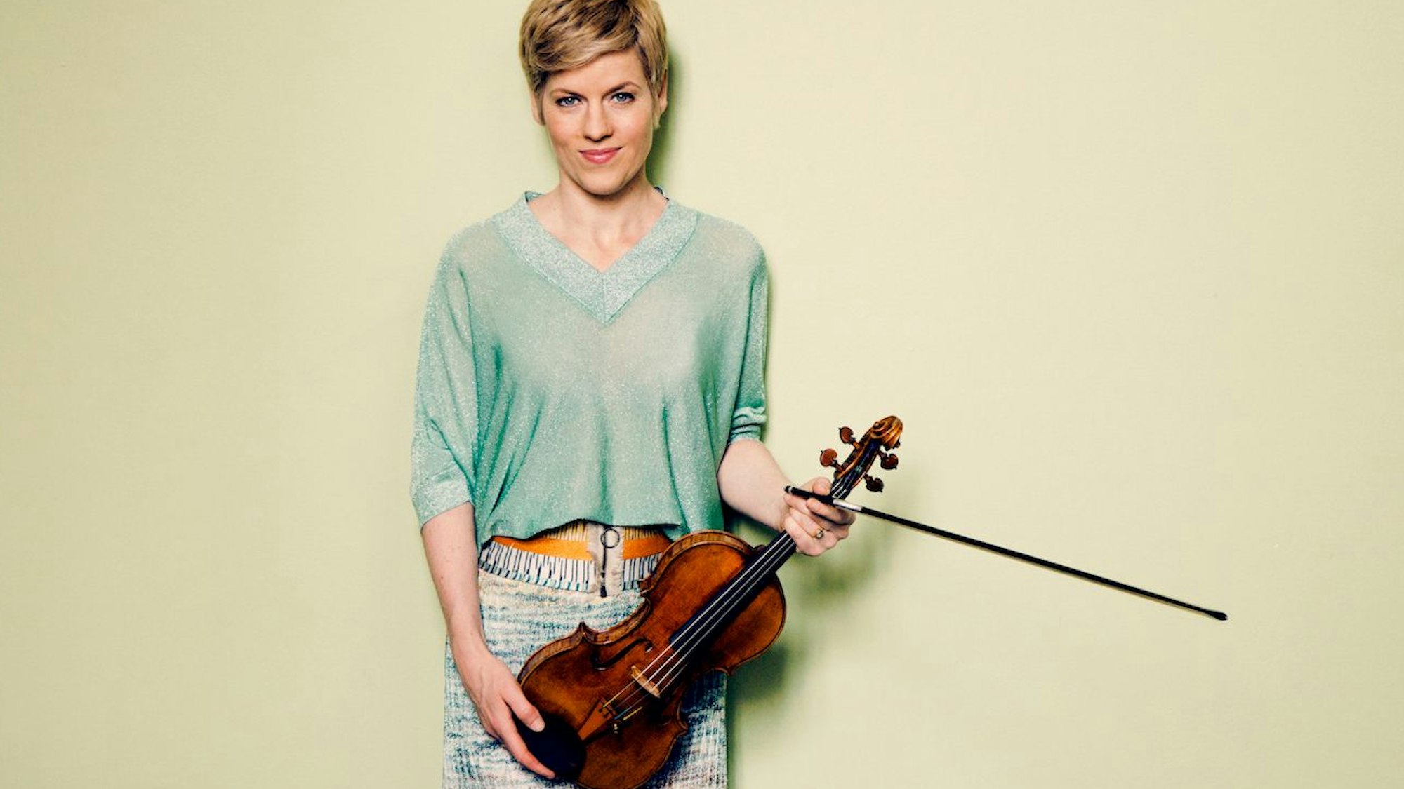 Isabelle Faust
hält Geige und Bogen in den Händen. Sie lächelt in die Kamera.
