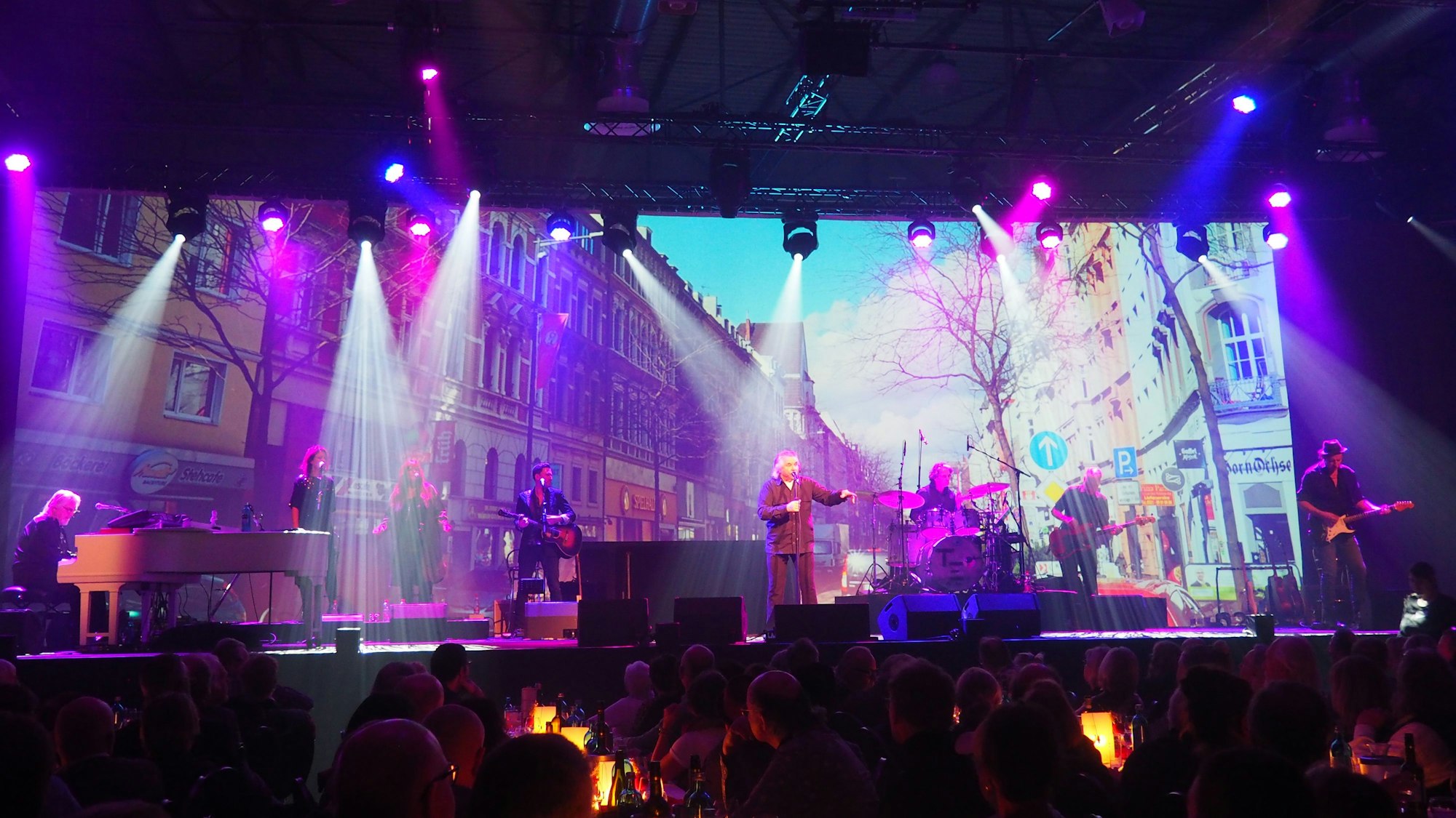 Tommy Engel und Band mit weißem Flügel und an Mikros vor einer Leinwand mit einem Bild einer Straße Kölns auf der Bühne.