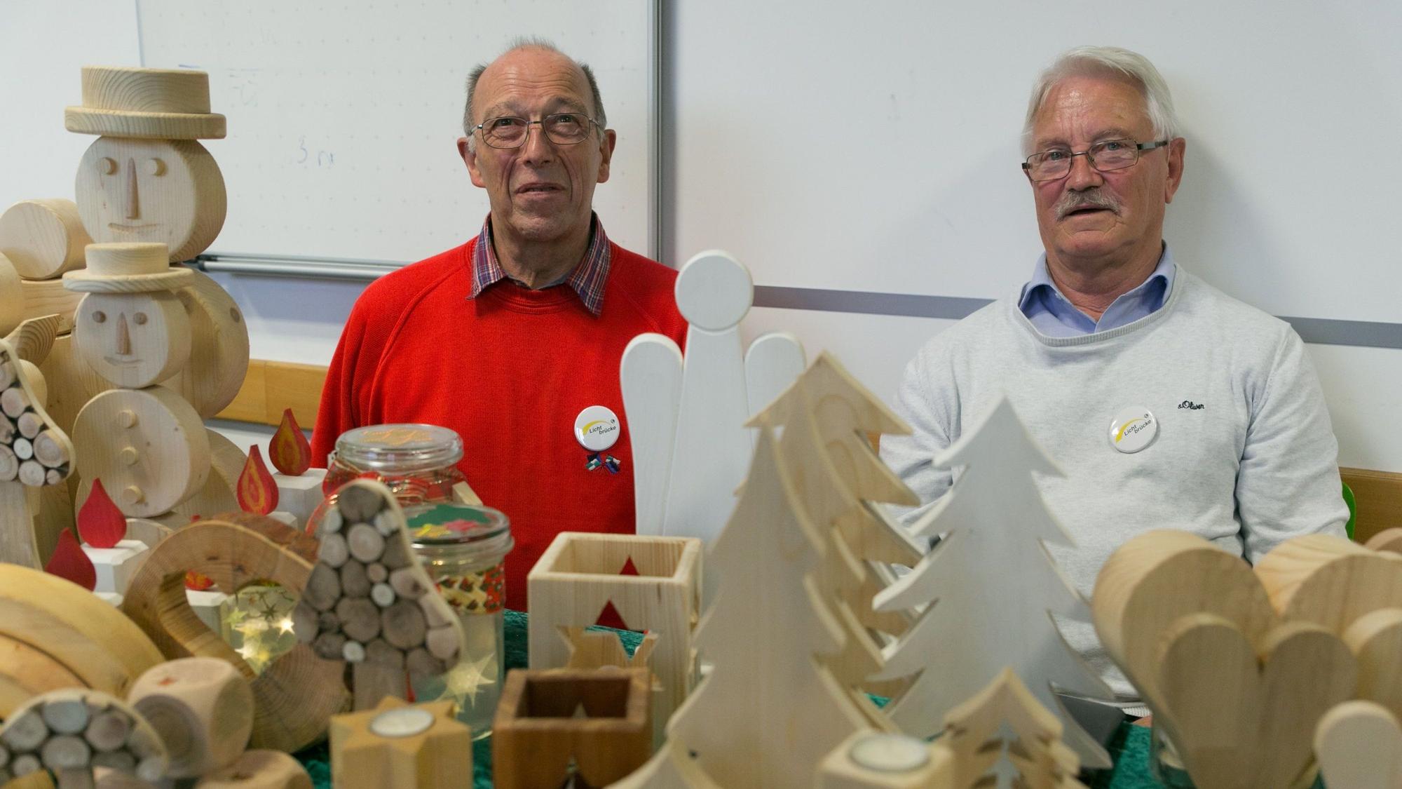 Gerhard Schmitz und Walter Schiffarth sitzen an ihrem Stand. Vor ihnen sind zahlreiche Holzschnitzereien aufgebaut.