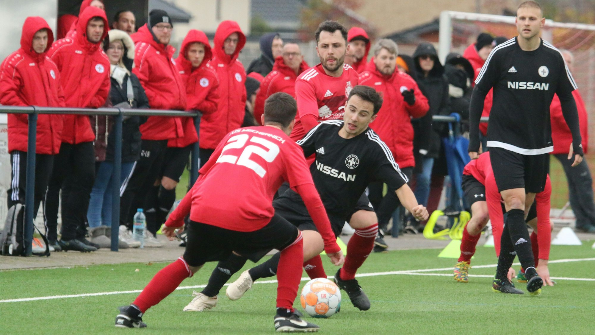 Ein Zülpicher Spieler gerät, bedrängt durch Mechernicher Gegner, ins Straucheln. Im Hintergrund stehen viele Zuschauer in knallroten Jacken des TuS Zülpich und haben die Kapuze über den Kopf gezogen.