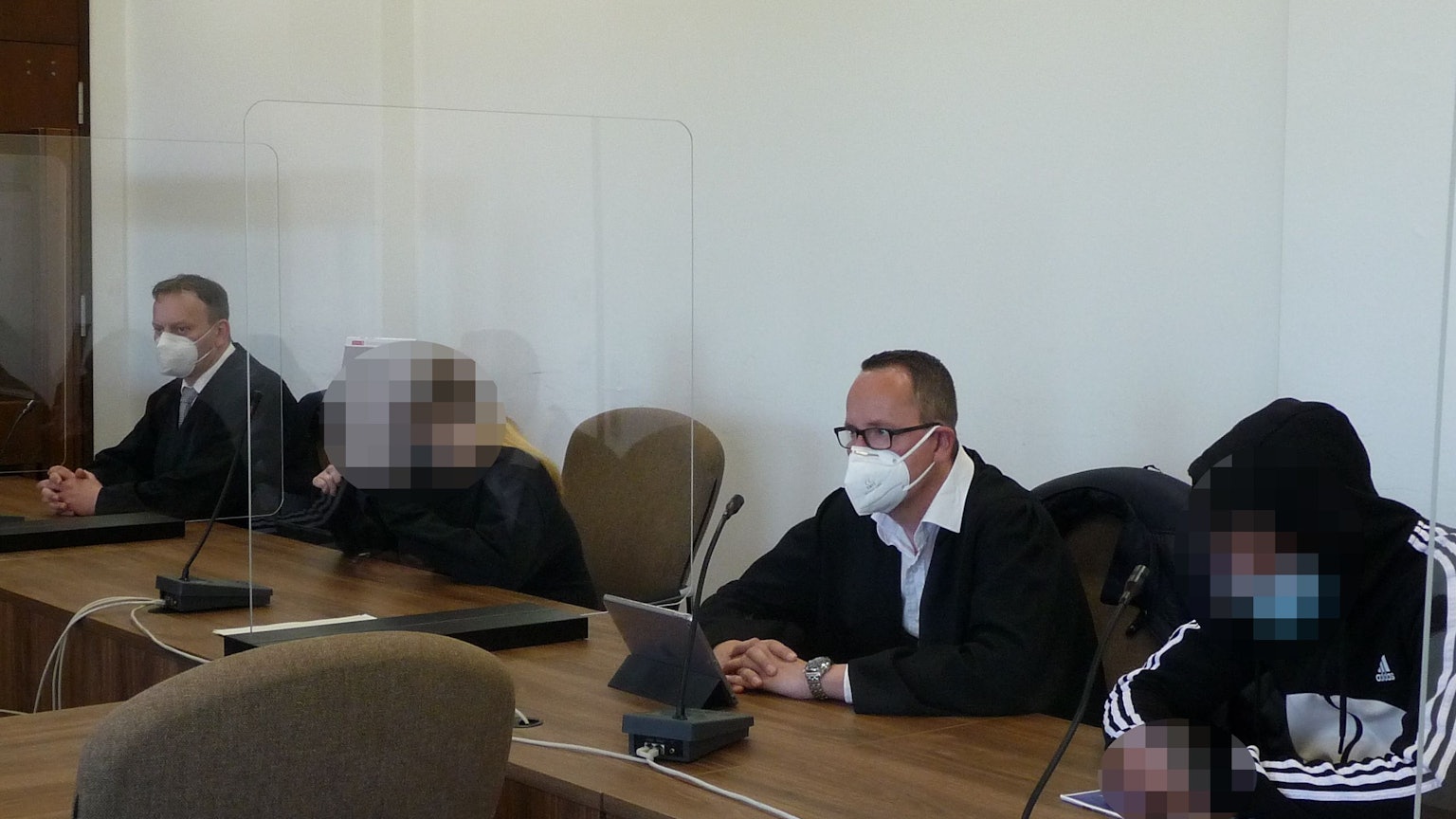 Eine Frau und ein Mann mit gepixelten Gesichtern sowie zwei Männer in Anwaltsrobe sitzen am Tisch einer Anklagebank im Kölner Landgericht.