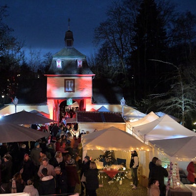 Ein Blick von oben auf den Weihnachtsmarkt an Schloss Eulenbroich in Rösrath. Besucher stehen an den zahlreichen, weißen Ständen. Im Hintergrund ist das angestrahlten Tor zum Schlossgelände zu sehen.