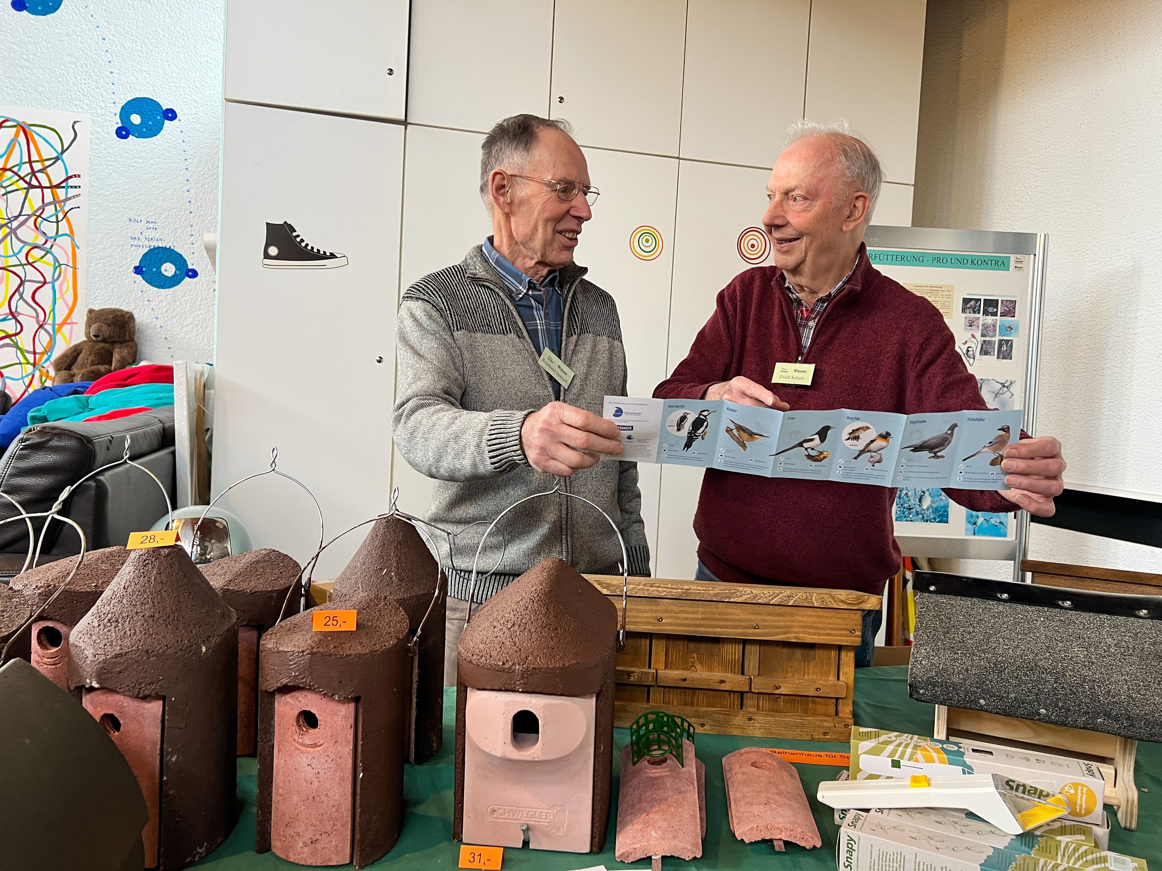 Zwei ältere Männer, Erich Schulz und Reinhard Dettlaff, stehen nebeneinander und schauen sich an. In der Hand halten sie ein Informationsblatt über Vögel.