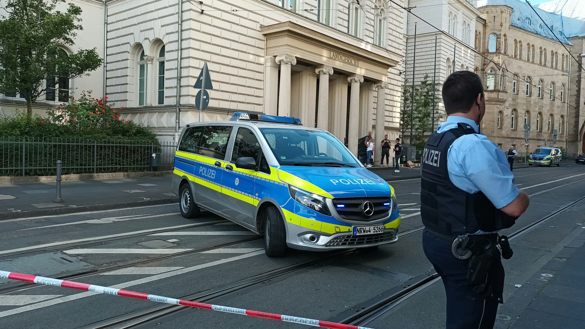 Ein Polizist und ein Streifenwagen stehen hinter einem Flatterband, mit dem der Zugang zum Landgericht Bonn abgesperrt ist.