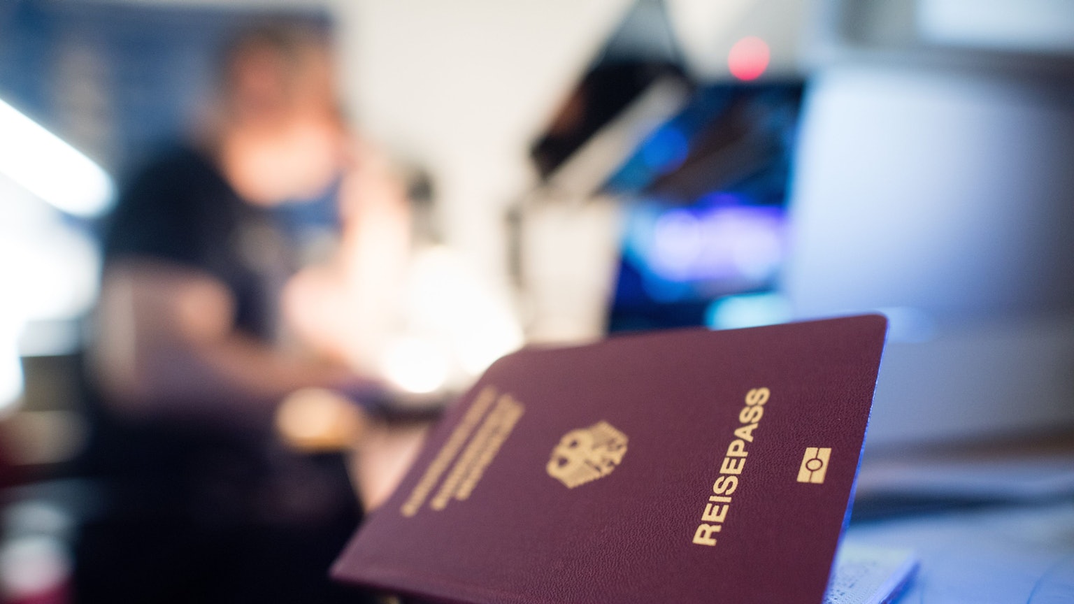 Ein deutscher Reisepass liegt auf einem Tisch, blaues Licht scheint darauf, eine Person ist verschwommen im Hintergrund zu sehen.