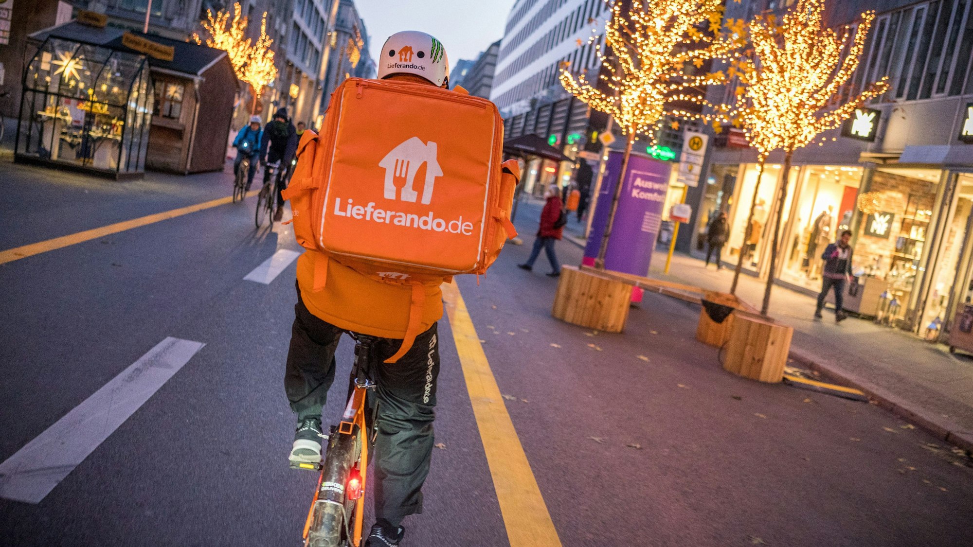 Ein Kurierfahrer des Restaurantlieferdiensts Lieferando fährt mit seinem Fahrrad über die Friedrichstraße in Mitte. Er hat einen Lieferando-Rucksack auf dem Rücken. Auf der Straße ist Weihnachtsbeleuchtung.