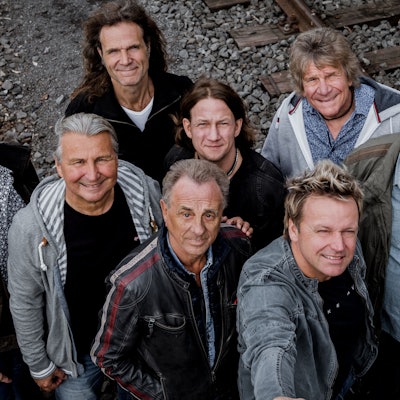 Die acht Bandmitglieder stehen auf einer Bahntrasse, von oben aufgenommen.