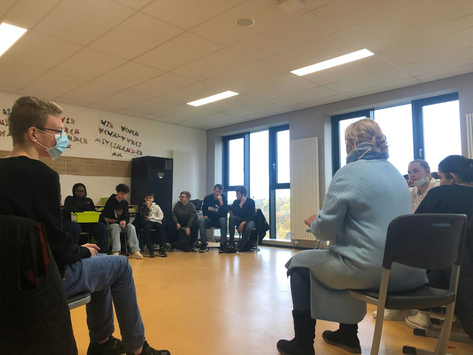 Eine erwachsene Frau sitzt auf einem Stuhl und hat den Rücken der Kamera zugewendet. Um sie herum sitzen im Stuhlkreis zahlreiche Schülerinnen und Schüler und sprechen mit ihr.