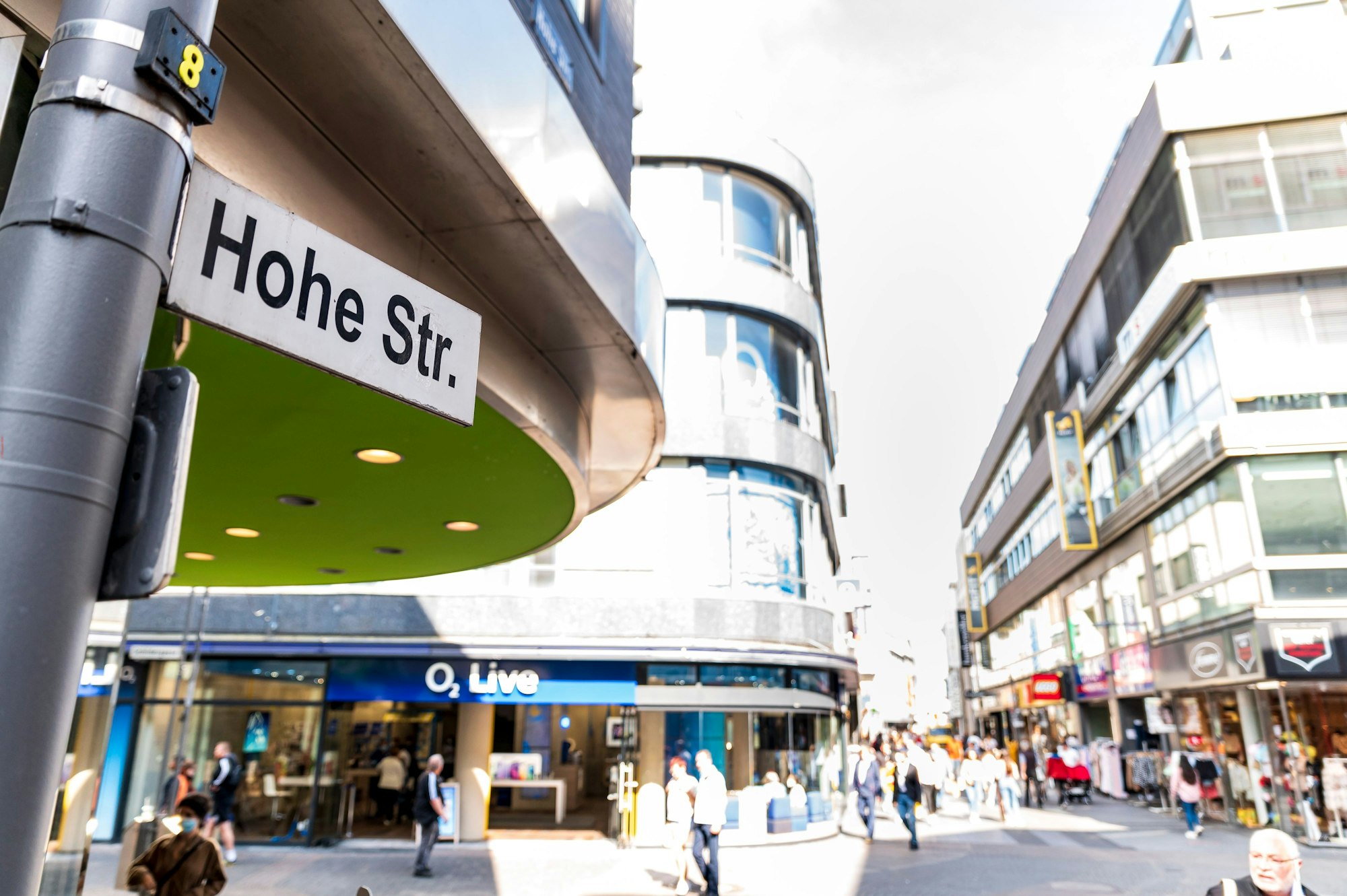 Das Bild zeigt ein Straßenschild mit der Aufschrift „Hohe Straße“, zusätzlich sind viele Passanten sowie Geschäftshäuser zu sehen.