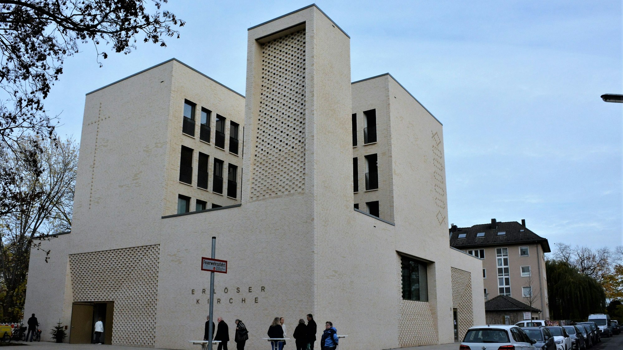 Die Erlöserkirche in Weidenpesch in Fassadenansicht: Heller Stein, kubisch verbaut und viele schmale Fenster sind zu sehen. Davor stehen Menschen im Gespräch.