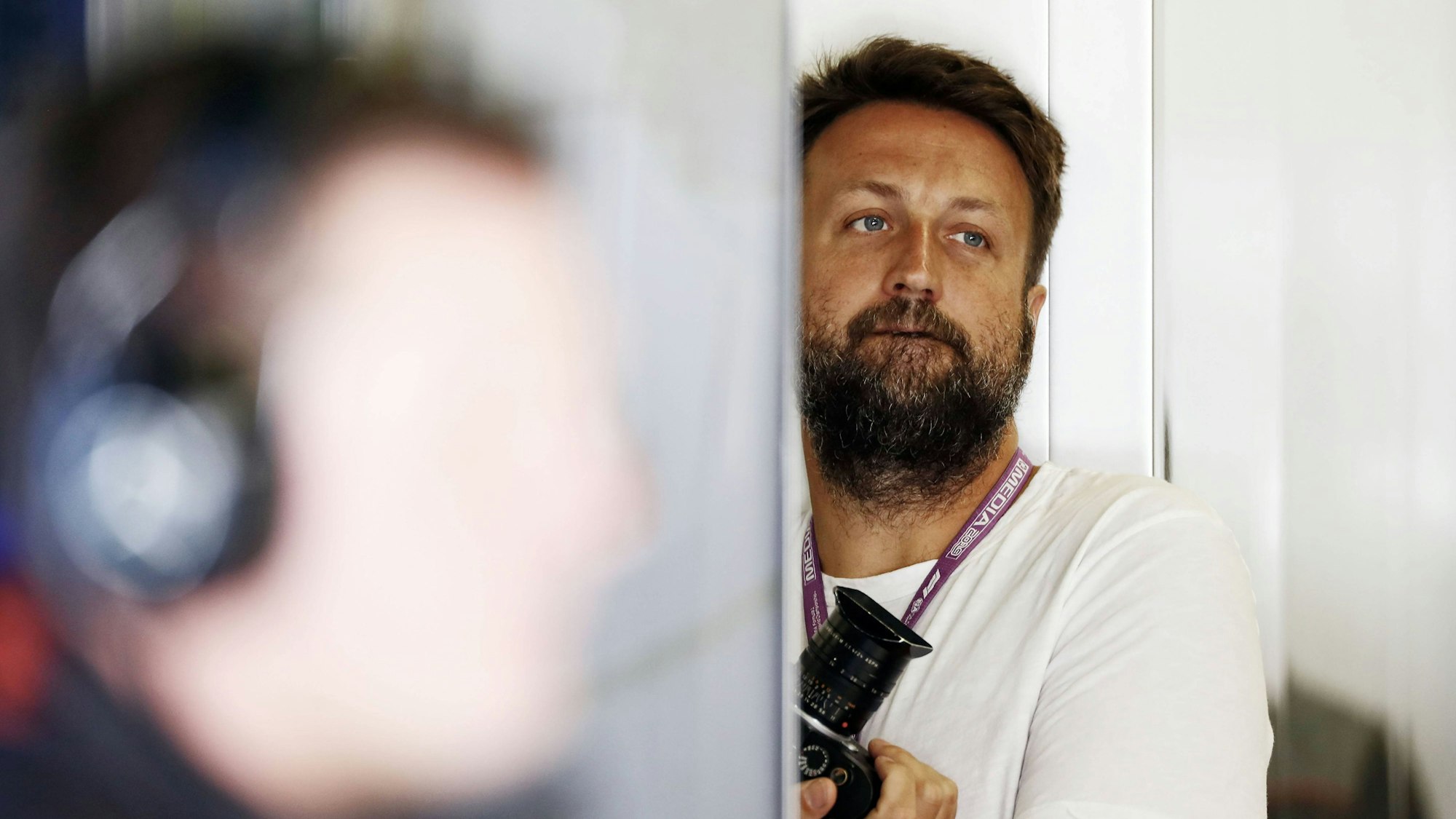 Der Fotograf Paul Ripke steht in weißem T-Shirt im Hintergrund des Bildes, er hält eine Kamera.