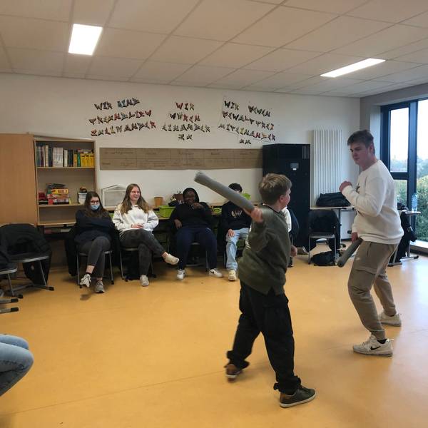 Ein großer und ein kleiner Junge kämpfen mit Schaumstoffstäben spaßeshalber miteinander. Vier Mitschülerinnen und Mitschüler schauen ihnen zu.