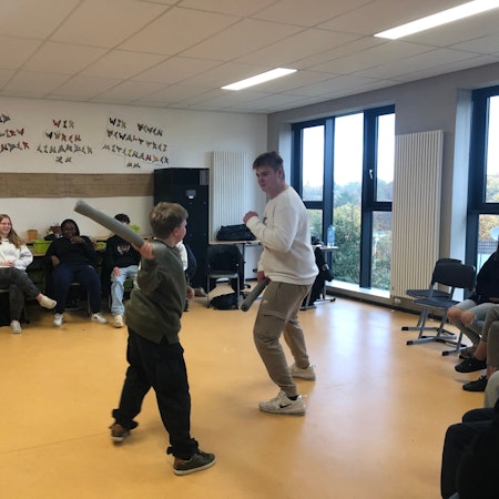 Ein großer und ein kleiner Junge kämpfen mit Schaumstoffstäben spaßeshalber miteinander. Vier Mitschülerinnen und Mitschüler schauen ihnen zu.