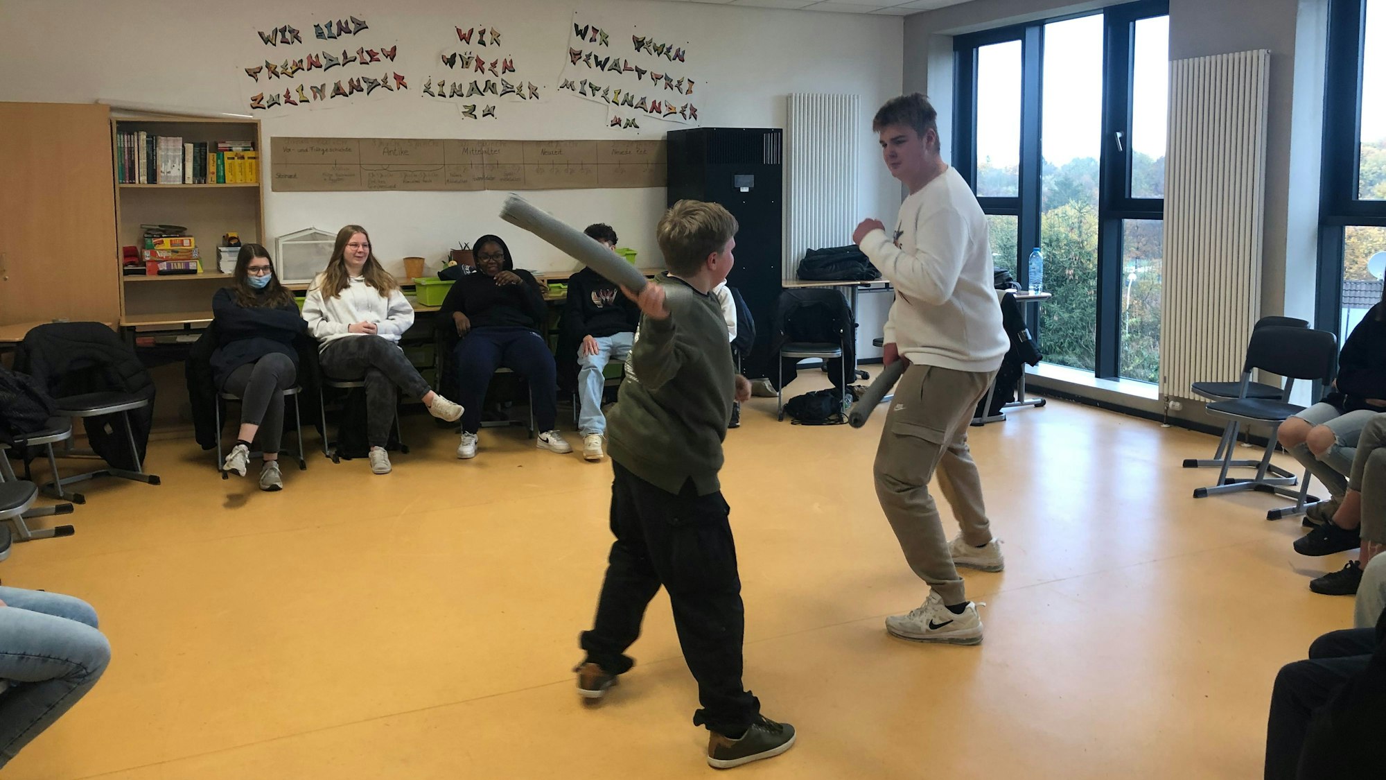 Zwei Schüler stellen mit Schaumstoffschlägern einen Kampf in der Mitte eines Stuhlkreises nach.