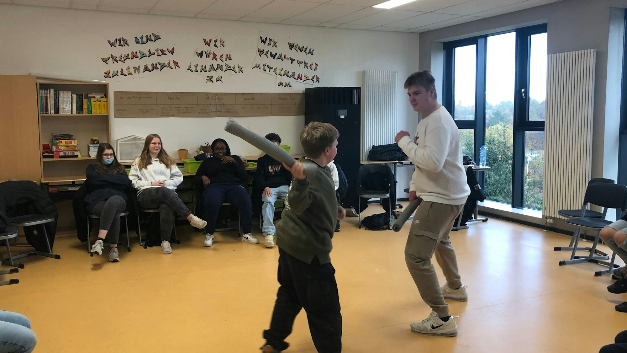Ein großer und ein kleiner Junge kämpfen mit Schaumstoffstäben spaßeshalber miteinander. Vier Mitschülerinnen und Mitschüler schauen ihnen zu.