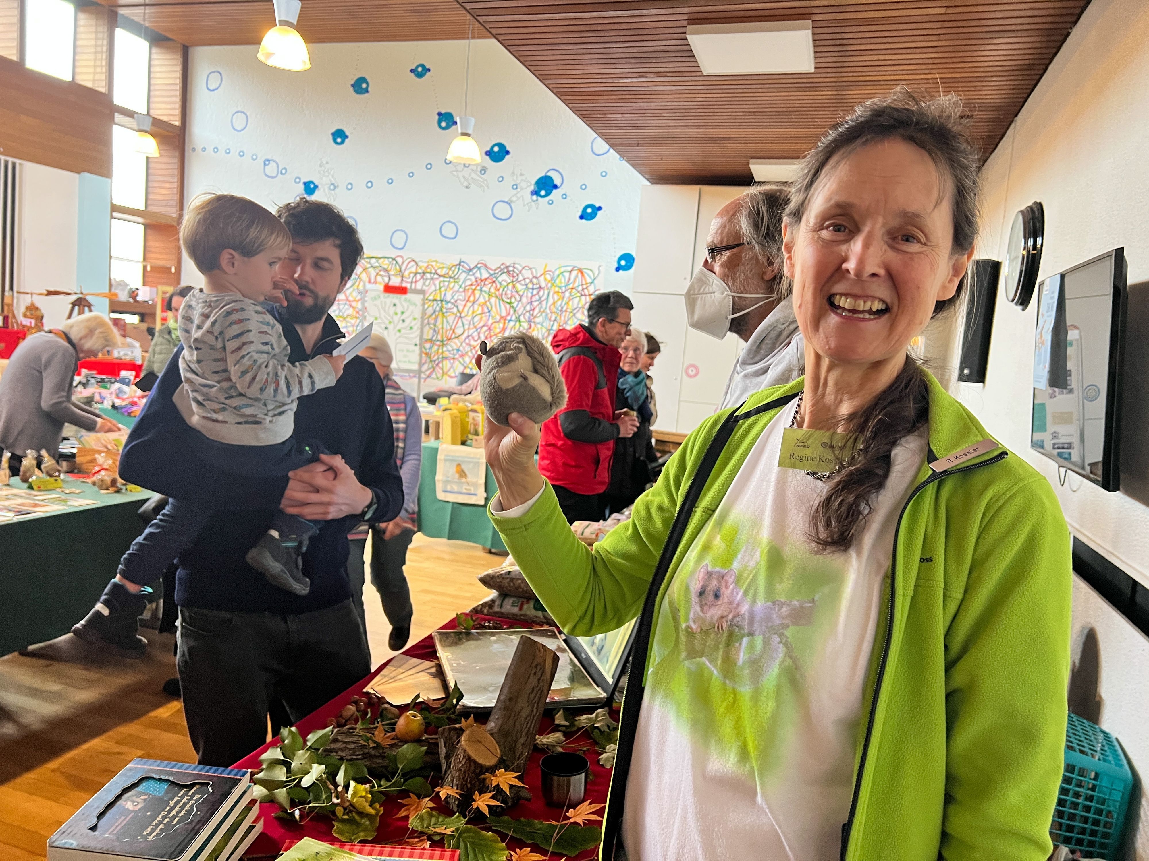 Eine Frau steht im Bildvordergrund, lächelt in die Kamera und hat ein Plüschtier in der Hand. Im Hintergrund sind Basarstände zu sehen und Menschen verschiedenen Geschlechts und Alters, die durch den Raum gehen.