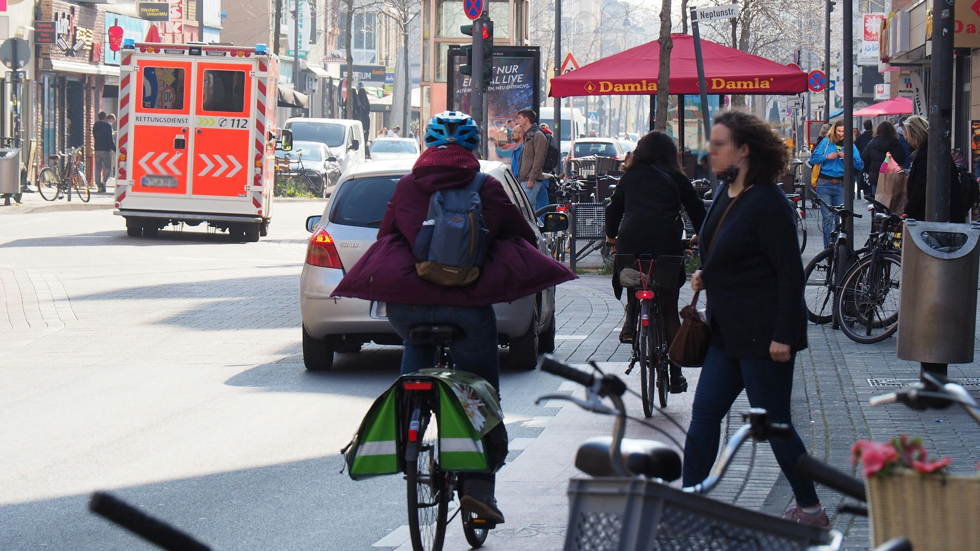 Unübersichtlicher Straßenverkehr auf der Venloer Straße. Ein Krankenwagen fährt auf der Straße, eine Fahrradfahrerin und eine Fußgängerin kreuzen sich.