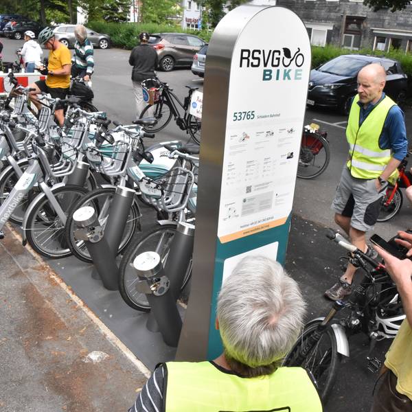 Menschen stehen um eine Stele der RSVG Bike-Vermietung herum. Dahinter sind E-Bikes aufgestellt.
