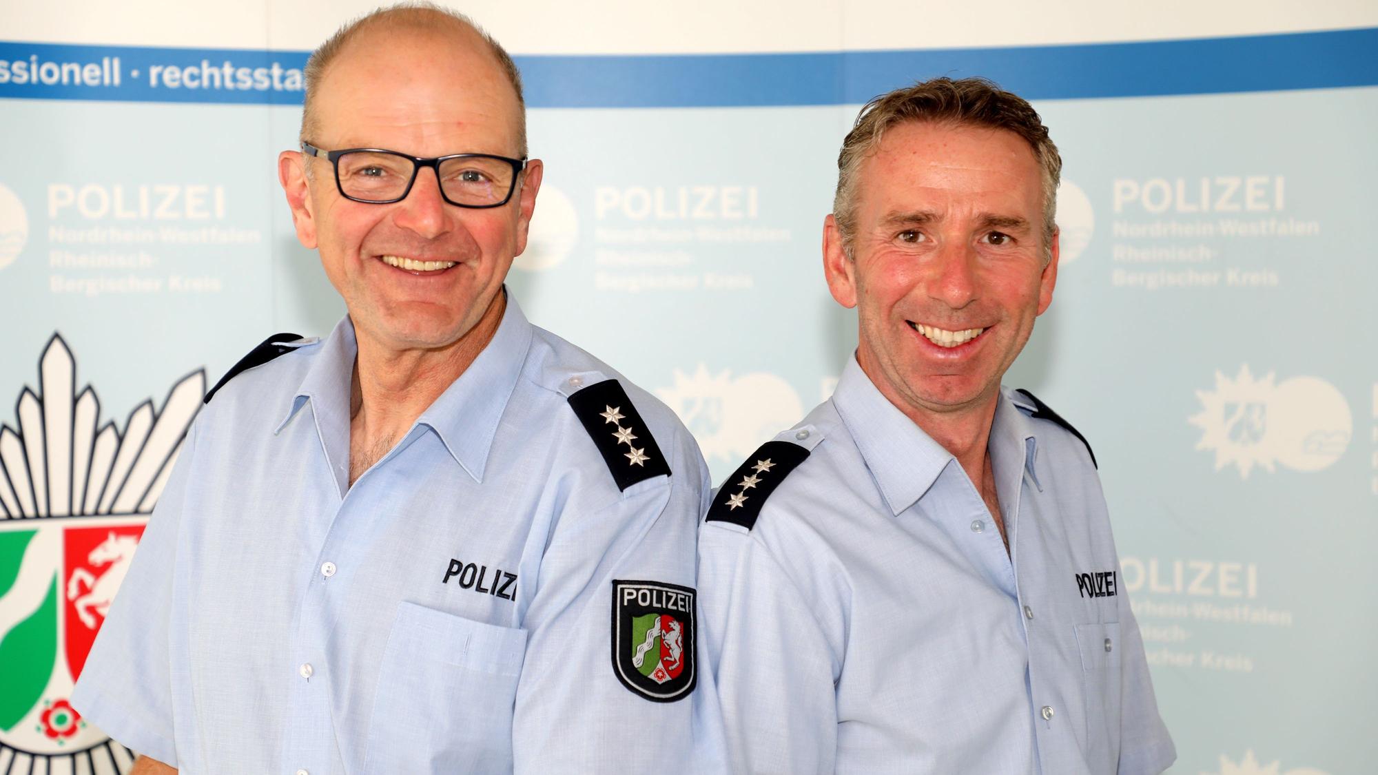 Peter Tilmans und sein Nachfolger Carsten Merkel stehen Schulter an Schulter vor einer Wand mit Polizeilogos darauf.