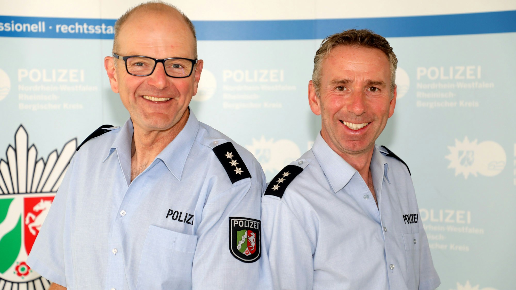 Peter Tilmans und sein Nachfolger Carsten Merkel stehen Schulter an Schulter vor einer Wand mit Polizeilogos darauf.