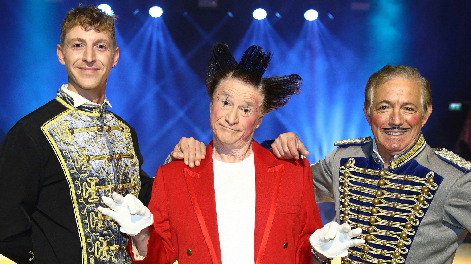 In der Mitte des Bildes steht Fumagalli-Clown Gianni Huesca im roten Sakko mit geschminktem Clownsgesicht und den charakteristischen, in drei Richtungen abstehenden Haaren. Links daneben steht sein Sohn Nico, rechts neben Fumagalli steht Bruder Daris.