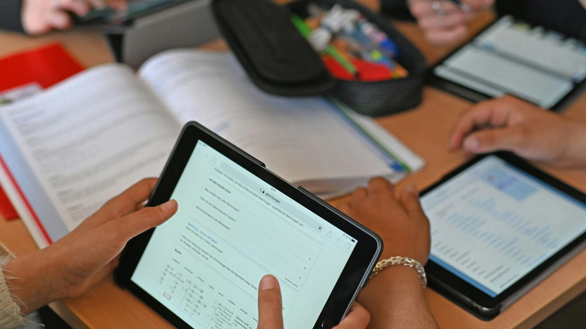 Auf einem Tisch liegen drei Tablets, auf denen getippt wird. Außerdem liegt ein Buch und ein Mäppchen mit Stiften auf dem Tisch.