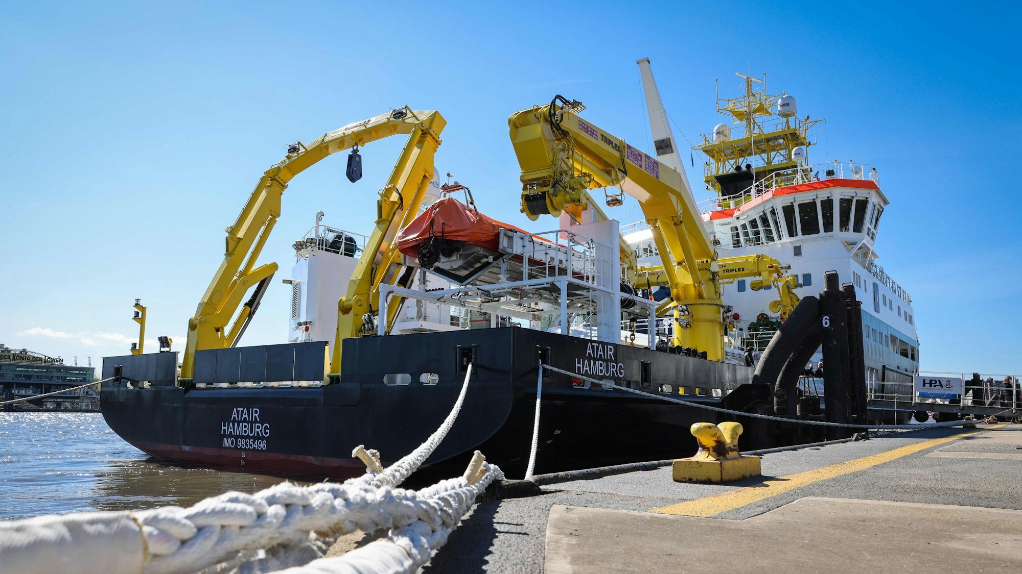 Das Schiff Atair liegt im Hamburger Hafen, es ist das Heck zu sehen. An Deck sind vor allem große gelbe Kräne sichtbar.