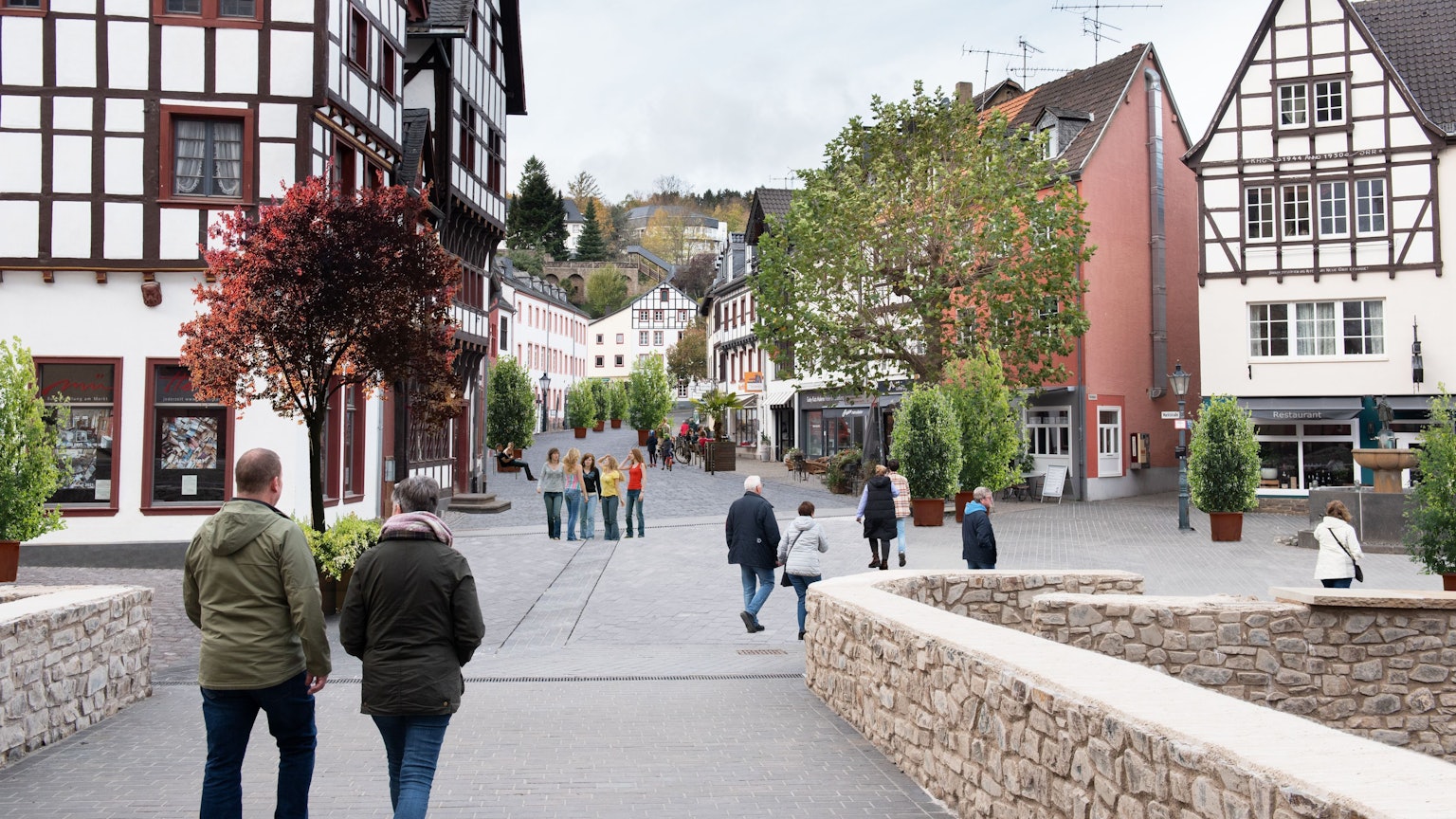 Das Bild zeigt den Marktplatz von Bad Münstereifel mit der Marktstraße im Hintergrund mit Blick von der Jesuitenkirche über die Heinz-Küpper-Brücke aus. Der Fotografie wurden Pflanzkübel und Bänke auf der Marktstraße hinzugefügt, die Autos wurden entfernt.