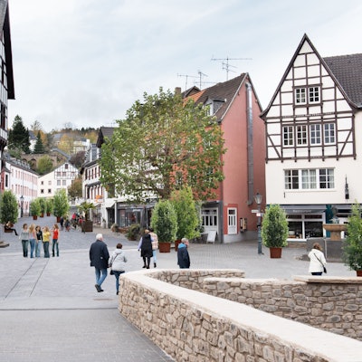 Das Bild zeigt den Marktplatz von Bad Münstereifel mit der Marktstraße im Hintergrund mit Blick von der Jesuitenkirche über die Heinz-Küpper-Brücke aus. Der Fotografie wurden Pflanzkübel und Bänke auf der Marktstraße hinzugefügt, die Autos wurden entfernt.