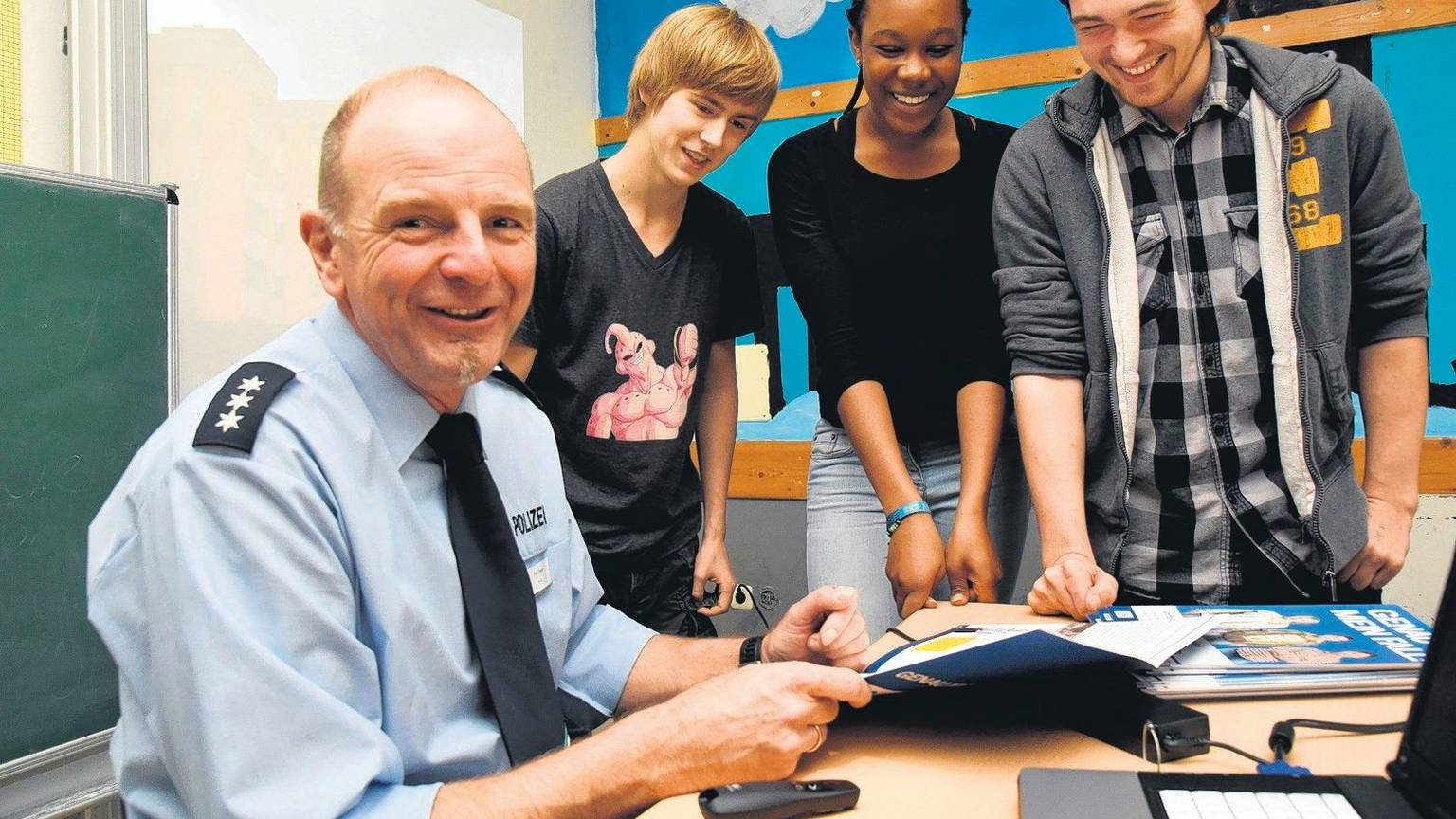 Peter Tilmans sitzt in Polizeiuniform an seinem Schreibtisch. Neben ihm stehen drei lächelnde Jugendliche.