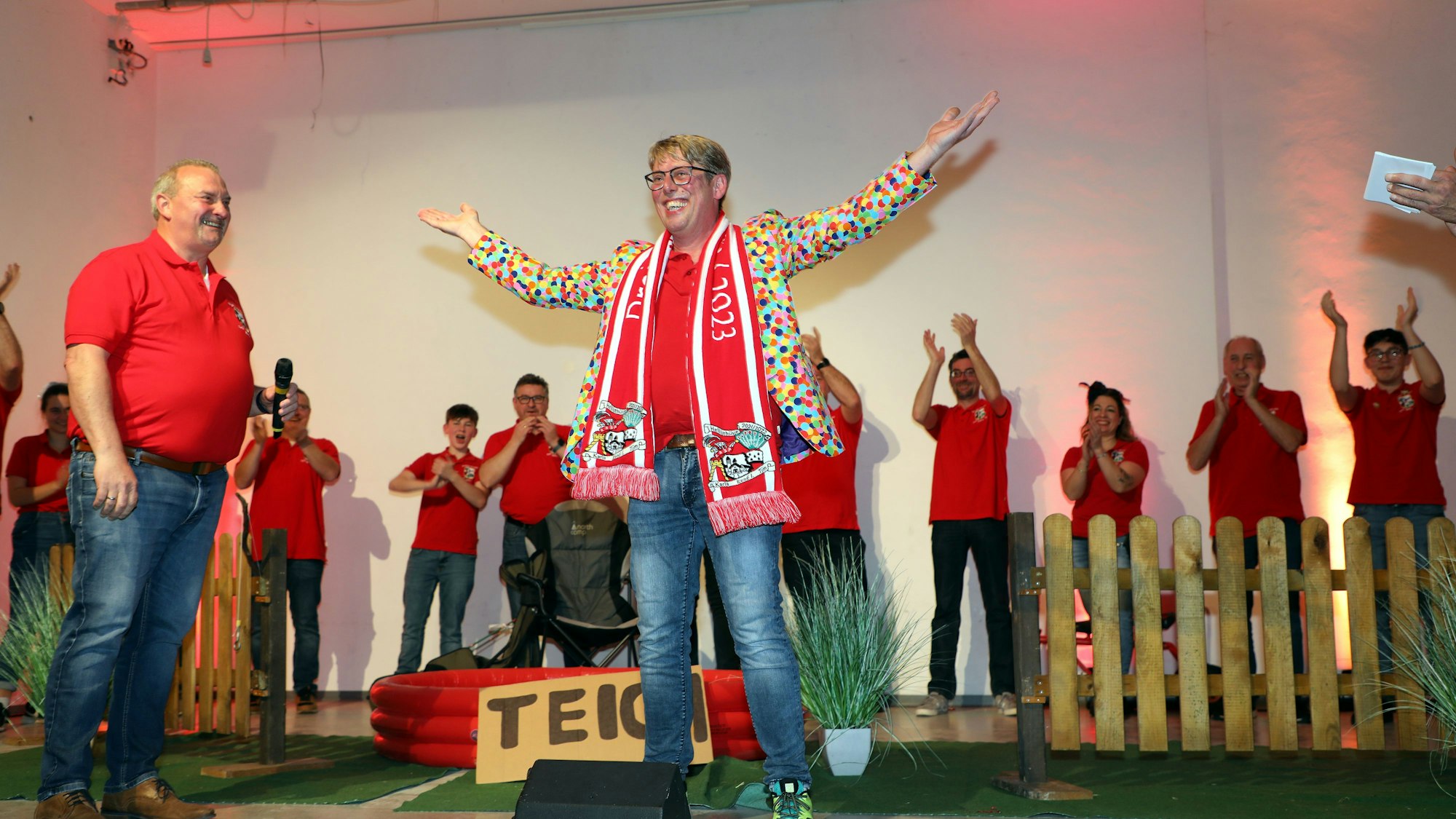 Prinz René I. (Leeser) steht mit Schal, Konfetti-Jacket und ausgebreitetem Armen auf der Bühne und strahlt. Neben und hinter ihm stehen in roten Shirts andere Heiligenhauser Karnevalisten.